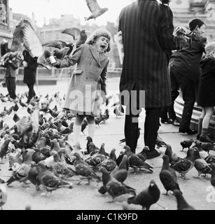 1950s. A young girl holding a tin of dried peas screams in terror as pigeons sit on her arm and surround her at Trafalgar Square, London, England, UK. Stock Photo