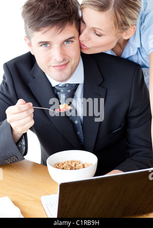 Smiling businessman eating breakfast using laptop while wife kissing his cheek Stock Photo