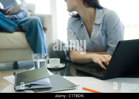 Woman using laptop in living room, looking over her shoulder at man sitting in background Stock Photo