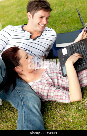 Couple of positive students using a laptop lying on the grass Stock Photo