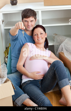 Happy couple expecting a baby sitting on the floor and holding the key of their new house Stock Photo