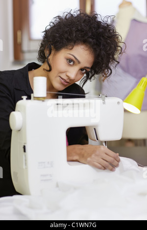 Young hispanic female dressmaker using sewing machine and smiling at camera. Vertical shape, side view, waist up Stock Photo