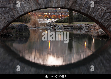 Gapstow Bridge as seen from the Pond with reflections in the water and a view of Wollman rink Stock Photo