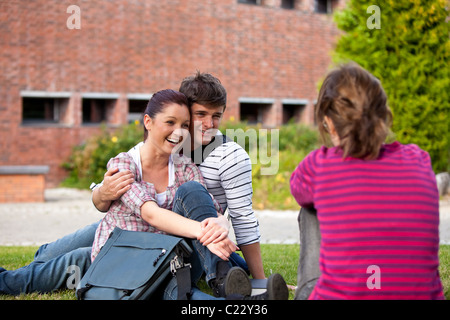 Happy couple of students sitting on grass talking with a female friend at their university campus Stock Photo
