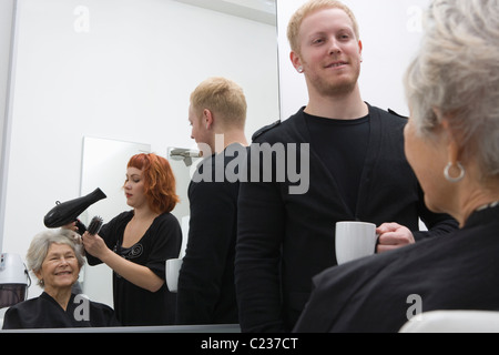 Stylist blow-drys elderly woman's hair:trainee stands with cup Stock Photo