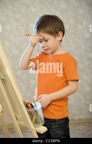 A young boy holding his hand with a pencil in up to his face:standing infront of a canvas with a pot of coloured pencils Stock Photo