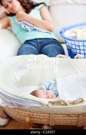Cute baby sleeping in his cradle with his mother lying on the couch in the background Stock Photo
