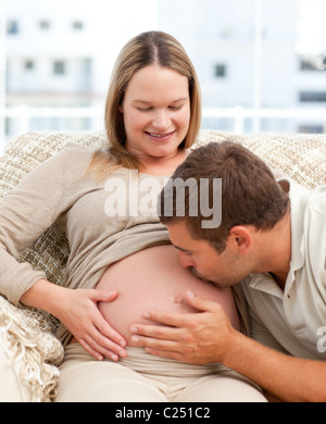 Proud man kissing the belly of his pregnant girlfriend sitting in the sofa Stock Photo