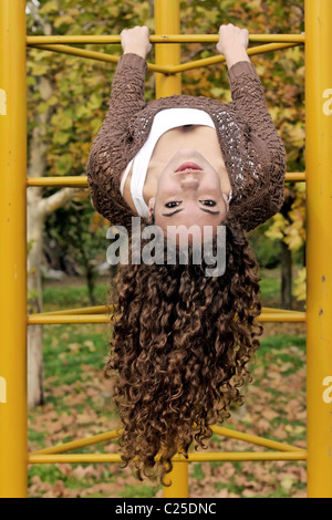 Curly brown haired girl hanging upside down in an outdoor playground Stock Photo