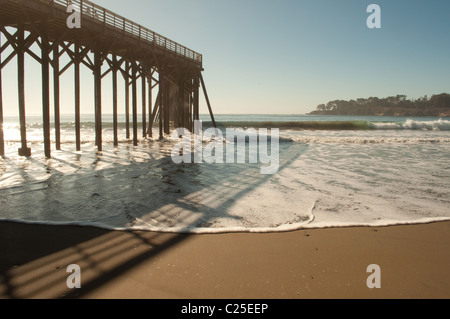 Wooden pier casts a shadow over foamy waves breaking against beach at low tide in San Simeon on the central California coast Stock Photo