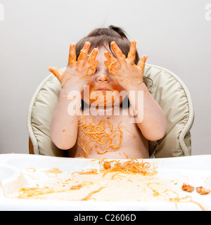 Happy baby having fun eating messy showing hands covered in Spaghetti Angel Hair Pasta red marinara tomato sauce. Stock Photo