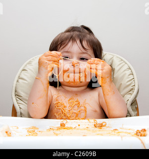 Happy baby having fun eating messy covered in Spaghetti holding Angel Hair Pasta red marinara tomato sauce.