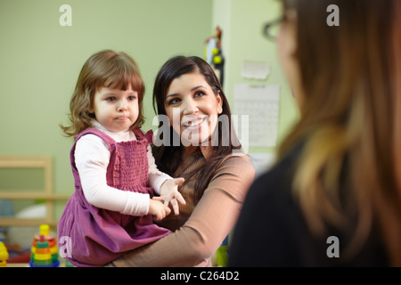 Caucasian female teacher and mom with baby girl talking in kindergarten. Horizontal shape, focus on background Stock Photo