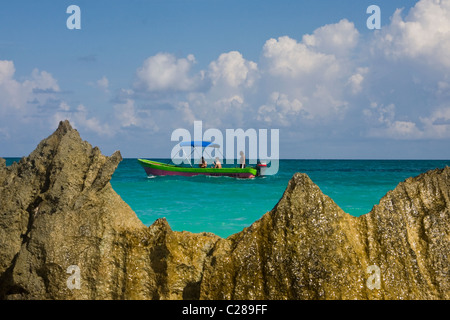 A colorful boat maneuvering in the Caribbean Sea with tourists and a rock formation in the foreground near la Playa Tulum Mexico Stock Photo