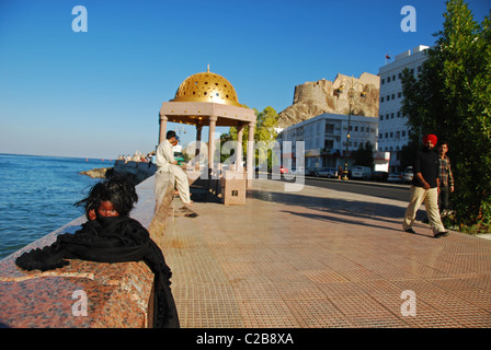Oman, Muscat, people sitting on parapet by sea with a doll's head placed in the foreground Stock Photo