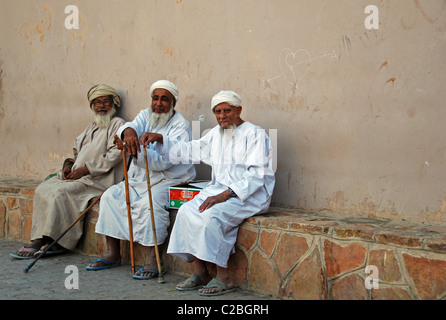 Oman, Nizwa, three senior men in traditional clothing sitting in front of a wall holding sticks Stock Photo
