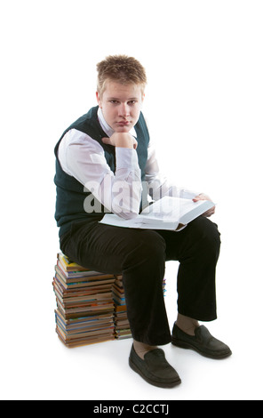 The schoolboy in a school uniform sits on a pack of books, with the opened book in hands Stock Photo