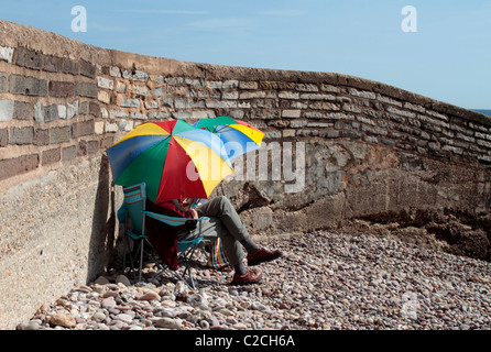 English couple sit on a beach in Devon using colorful sunshades Stock Photo