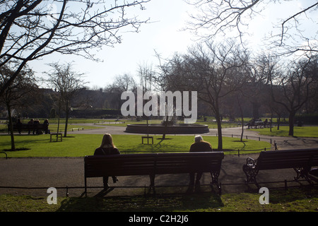 Two people, an old man and a young girl sitting on a park bench in sunshine in St Stephens Green, Dublin, Ireland. Stock Photo