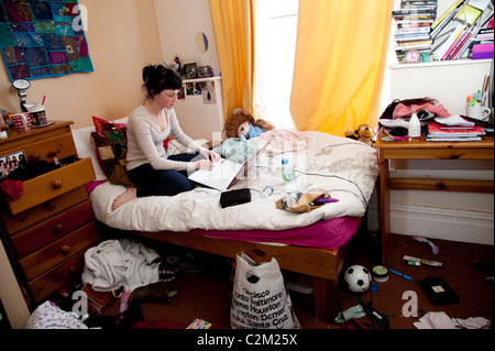 A young woman UK university student working on her laptop computer on the bed in an untidy bedroom Stock Photo