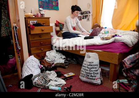 A young woman UK aberystwyth university student working on her laptop computer on the bed in an untidy bedroom Stock Photo