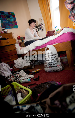 A young woman UK aberystwyth university student working on her laptop computer on the bed in an untidy bedroom Stock Photo