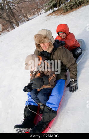 Boy and girl ages 6 and 7 sliding down snowy winter Merriam Park hill with dad age 42. St Paul Minnesota MN USA Stock Photo