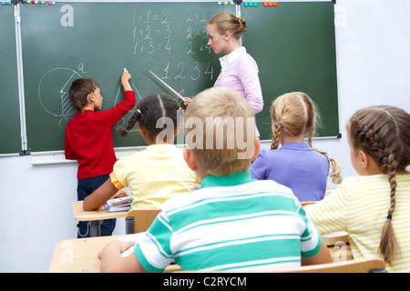 Photo of elementary student by blackboard looking at his teacher helping him Stock Photo
