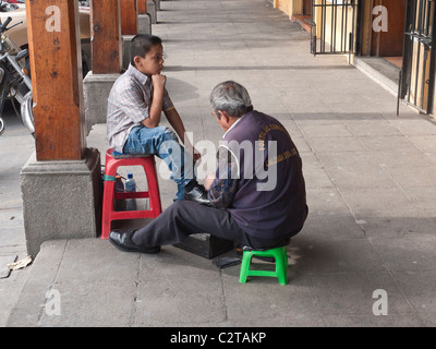 A young boy gets his shoes shined by an older adult shoe shine man in an open street side arcade in Antigua, Guatemala. Stock Photo