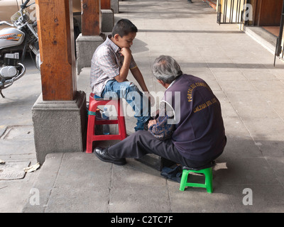 A young boy gets his shoes shined by an older adult shoe shine man in an open street side arcade in Antigua, Guatemala. Stock Photo