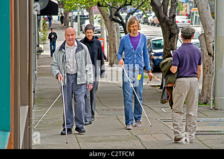 Accompanied by an instructor, a blind man using a white cane teaches two young volunteers how to assist handicapped people on Fi Stock Photo