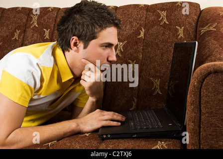 Young man lying on couch in living room using laptop and holding hand to chin thinking Stock Photo