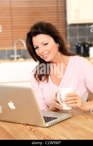 Woman using her laptop in the kitchen holding a mug of tea or coffee Stock Photo