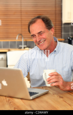 Happy mature man using his laptop in the kitchen holding a mug of tea or coffee Stock Photo