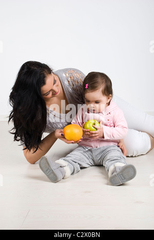 Young mother sitting on wooden floor with her baby girl and giving her child fruits Stock Photo
