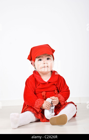 Little girl 11 months old sitting on floor dressed in little red redding hood costume and holding a shoe in her hands Stock Photo