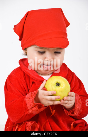 Baby girl in red clothes holding an apple in her hands and looking at fruit with lust and tongue out like she say Stock Photo