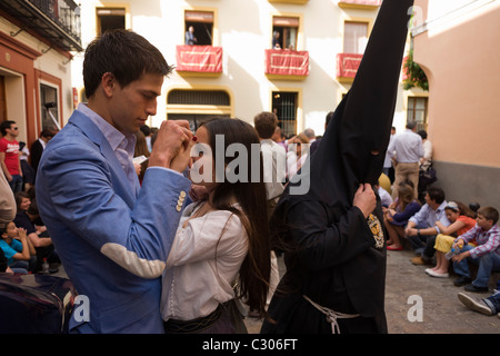 Hooded penitents (Nazarenos) walk past young lovers during Seville's annual Easter Holy Week Stock Photo