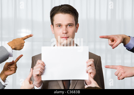 Serious businessman with blank paper looking at camera surrounded by pointing hands Stock Photo