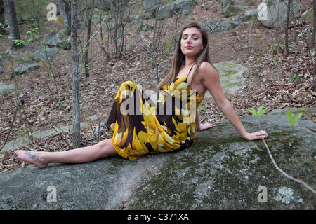 Pretty woman in a yellow dress sitting on a rock in the woods with thoughtful and friendly eye contact Stock Photo