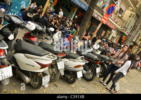Horizontal wide angle of disorganised chaos on the streets of Hanoi with mopeds & motorbikes parked everywhere whilst people eat Stock Photo