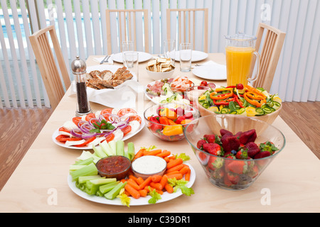 Dining table laid for a healthy lunch of fresh salad, vegetables, fruit, bread, ham, prawns, olives, strawberries & orange juice Stock Photo