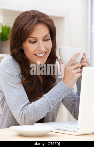 Beautiful brunette woman at home sitting at a table using her laptop computer, smiling and drinking tea or coffee Stock Photo