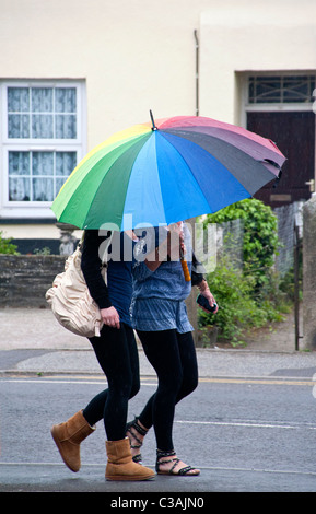 Two young women friends sharing a large, colourful / colorful umbrella during a rain shower. Neither of them are recognisable Stock Photo