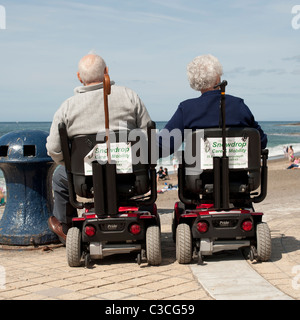 Rear view of an elderly disabled couple husband and wife at the seaside sitting in powered mobility scooters, Aberystwyth Wales Stock Photo