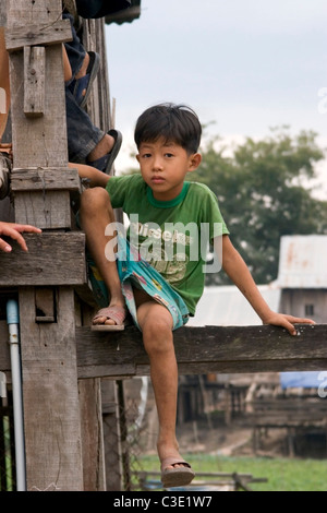 A young boy living in poverty is sitting on his rundown wooden home on a dirt street in Kratie, Cambodia. Stock Photo