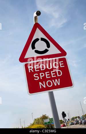 Reduce speed now sign for roundabout in England. Showing traffic and lights in distance. Shot on sunny day. Blue sky. Stock Photo