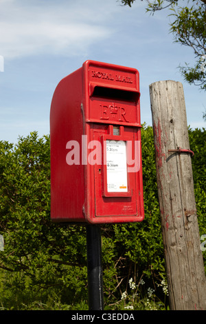 Royal mail post box on road side in rural setting. Showing small red post box with trees, shrubs blue sky clouds. Sunny day. Stock Photo