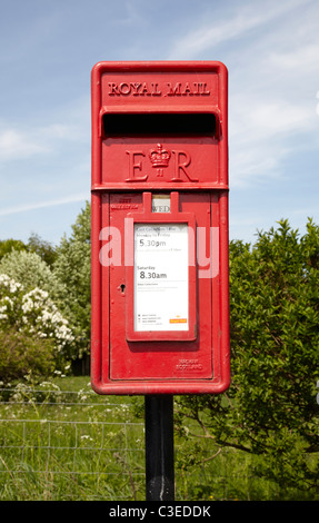 Royal mail post box on road side in rural setting. Showing small red post box with trees, shrubs blue sky clouds. Sunny day. Stock Photo