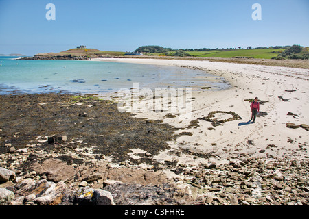 Woman crossing sandy Old Grimsby harbour beach on Tresco in the Isles of Scilly with the Old Blockhouse on the headland Stock Photo